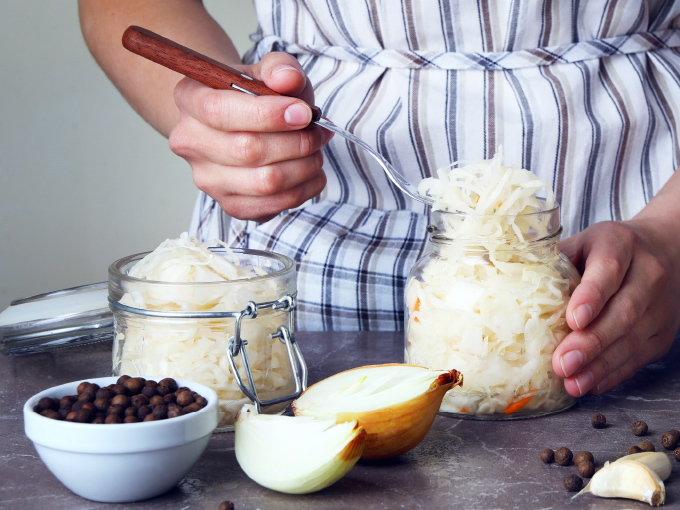 Hände einer Frau halten ein Glas mit Sauerkraut auf einem grauen Tisch, umgeben von Zutaten.