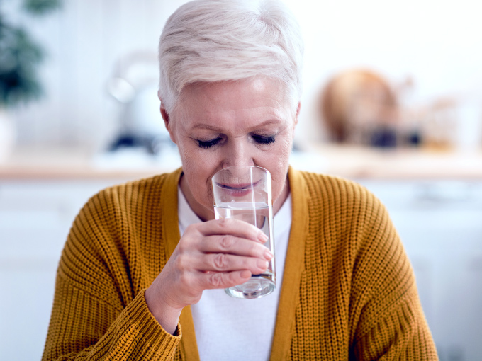 Eine ältere Frau trinkt ein Glas Wasser in ihrer Wohnung